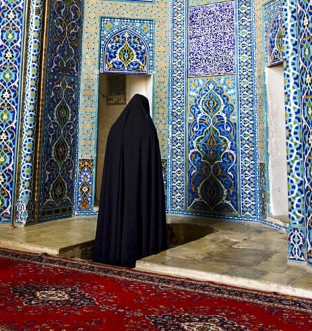 A person in a black cloak stands in a small niche within an ornately decorated room with intricate blue and white tilework and a red carpet, capturing the warmth of Iran's beauty.