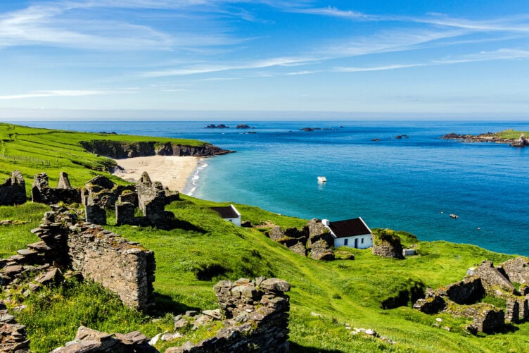 Explore the picturesque coastal landscape of Ireland, where ancient stone ruins meet lush green fields overlooking a sandy beach and the shimmering blue sea under a clear sky.
