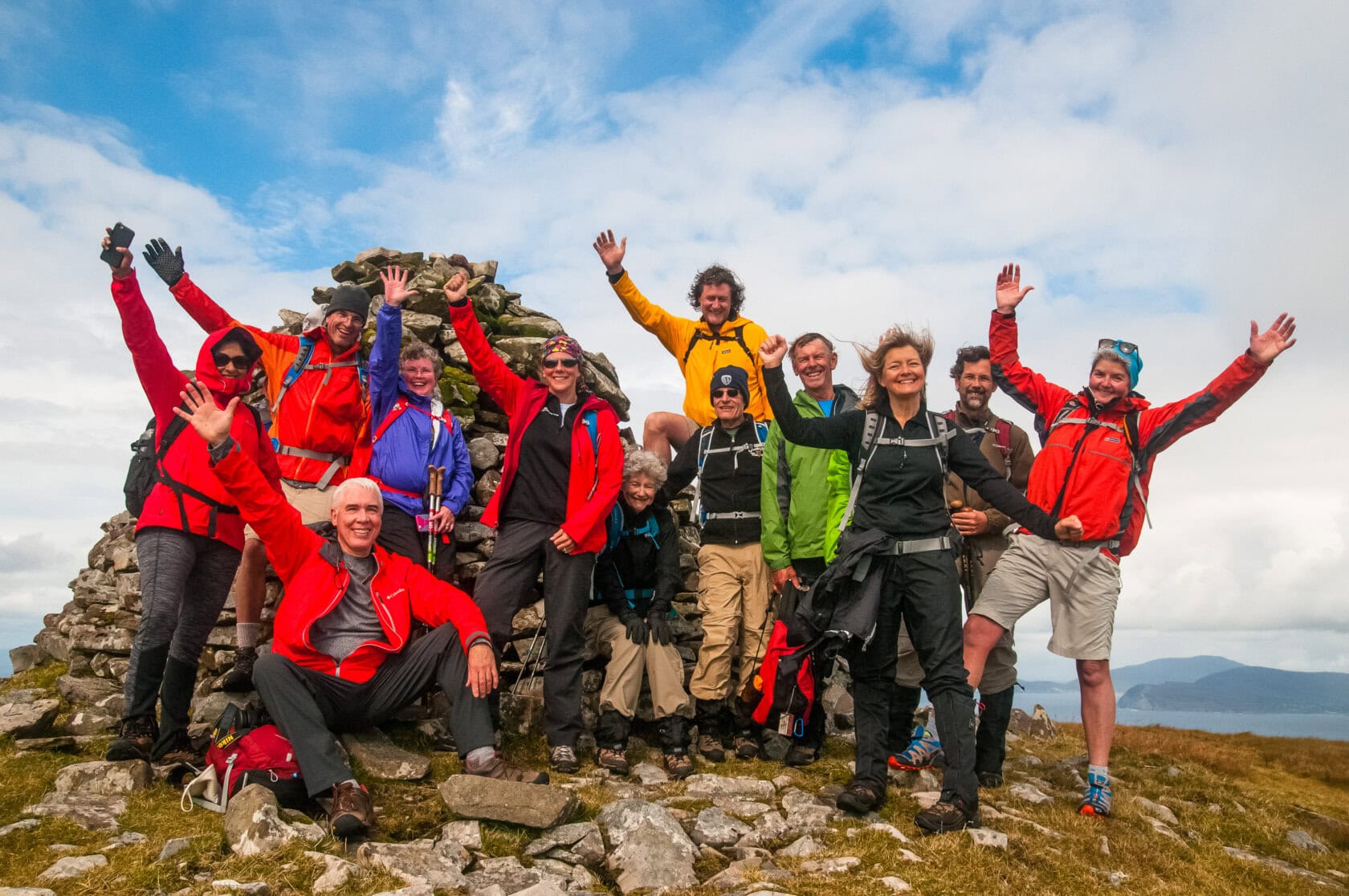 Group of people in hiking attire posing and raising arms near a stone cairn on a rocky landscape with a cloudy sky, enjoying one of the top 11 things to do in Ireland.