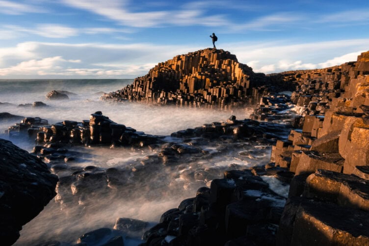 A traveler stands on the basalt columns at Giant's Causeway, surrounded by misty waves under a blue sky with scattered clouds—a quintessential Ireland experience for any travel enthusiast.