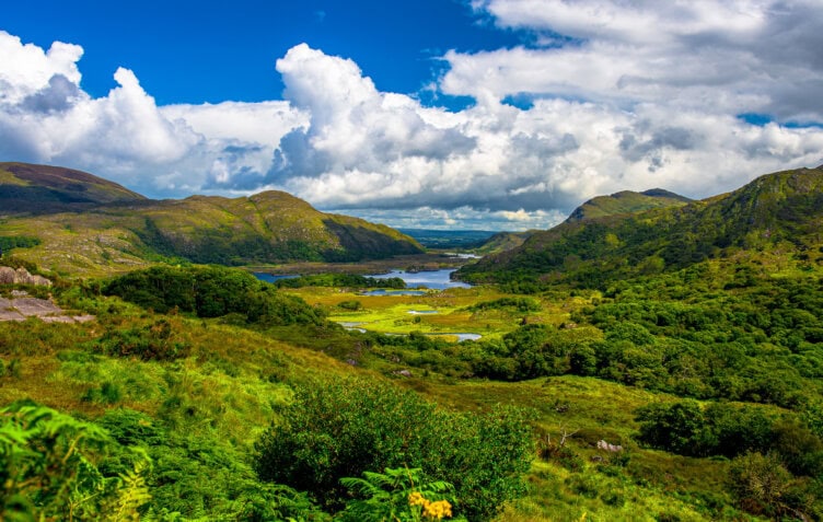 Explore the lush green valley of Ireland, where a river gently flows through, surrounded by majestic mountains under a partly cloudy sky—an ideal travel destination with endless things to do.