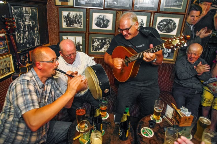 Musicians play flute, drum, and guitar in a lively Irish pub with people clapping. Tables have drinks, and the walls are adorned with framed photos. A must-do for anyone traveling to experience Ireland's vibrant culture.