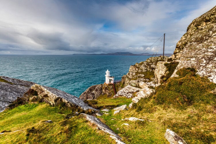 Explore the enchanting lighthouse on Ireland's rocky coast, nestled amidst lush green grass and offering breathtaking views of a vast ocean under a cloudy sky. A must-see in your list of things to do.
