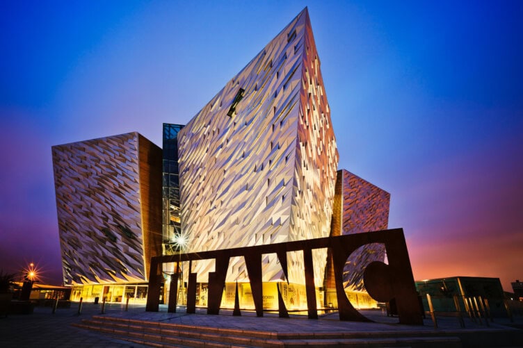 A modern angular building in Ireland glows at sunset, its facade illuminated beneath a large "TITANIC" sign. Ranked among the Top 11 must-see sights, it's one of the top things to do for visitors seeking history and architecture.