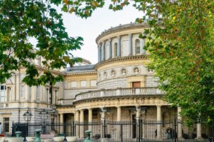 In Dublin, a historic stone building with large round towers and arched windows peeks through the trees, surrounded by a metal fence—a must-see for any travel itinerary.