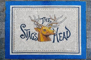A mosaic artwork depicting a stag's head with "The Stags Head" text is framed by a blue border on a tiled surface, adding charm to one of Dublin's unique spots. Perfect for those exploring the city's vibrant art scene and seeking memorable things to do.