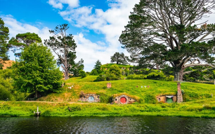 Experience one of New Zealand's top 15 attractions: a scenic view of hobbit-style houses nestled into grassy hills, surrounded by large trees and a shimmering pond in the foreground, under a partly cloudy sky. It's an enchanting destination with so many things to do and explore.