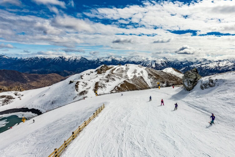 Skiers navigate a snowy mountain slope under a partly cloudy sky, with New Zealand's rugged mountain range visible in the background, offering a stunning scene straight out of a travel guide.