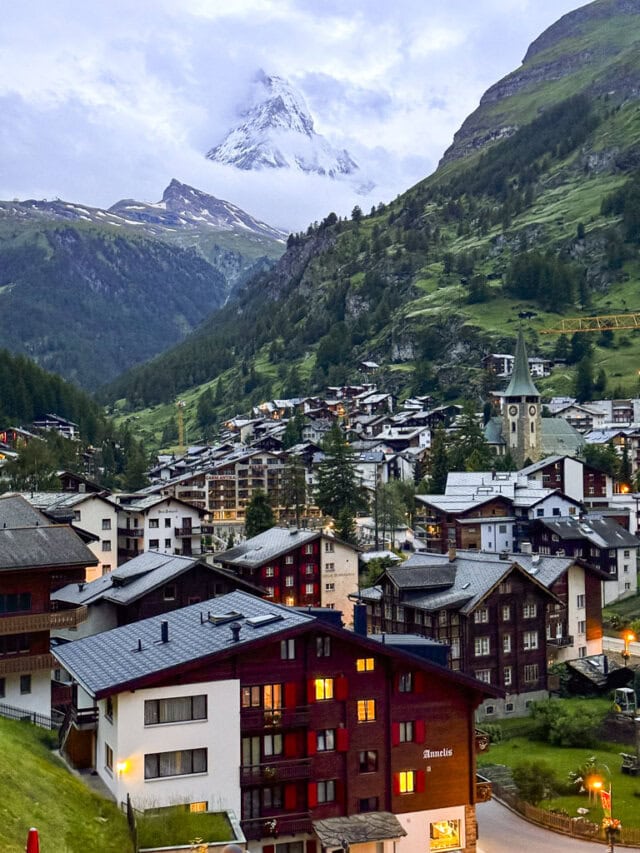Mountain village nestled in a valley, with wooden houses, a prominent clock tower, and the majestic Monte Rosa towering snow-capped in the distance—a perfect spot for those planning to embark on the Tour de Monte Rosa hiking adventure.