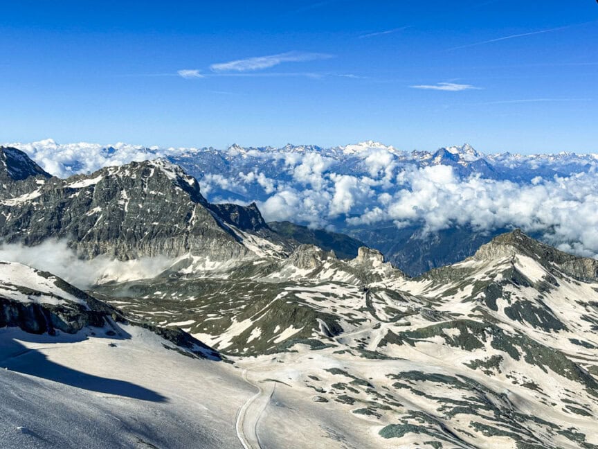 The snow-covered mountain range under a blue sky with scattered clouds offers breathtaking views. Jagged peaks and valleys, reminiscent of Monte Rosa's dramatic terrain, beckon adventurous hikers exploring the Tour de Monte Rosa trail in the distance.