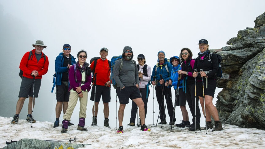 A group of ten hikers, equipped with walking poles, stands on the snowy and rocky terrain of the Tour de Monte Rosa. The foggy backdrop reveals jagged rocks and scattered patches of snow, encapsulating the true essence of alpine hiking adventure.