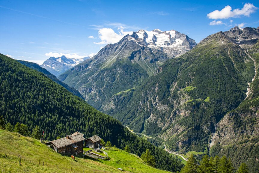 Rustic cabin on a grassy hillside with lush green valleys and towering snow-capped mountains under a clear blue sky offers the perfect respite after an exhilarating day of hiking amid the breathtaking scenery of Tour de Monte Rosa.