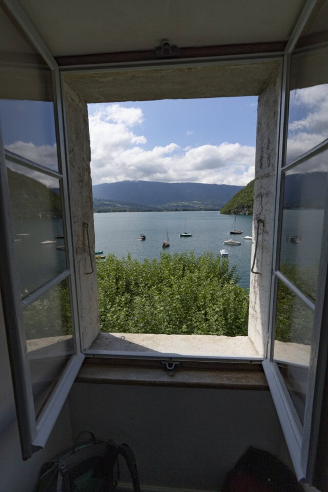 View from an open window overlooking a lake with several sailboats and surrounding hills, perfect for hiking enthusiasts exploring scenic trails under a cloudy sky.
