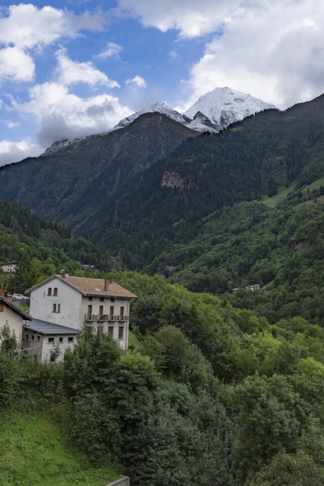 A white building with a red roof is nestled among dense green trees, set against the backdrop of tall mountains reminiscent of Mont Blanc, under a partly cloudy sky. The scene invites thoughts of hiking through serene paths amidst an autumnal landscape.
