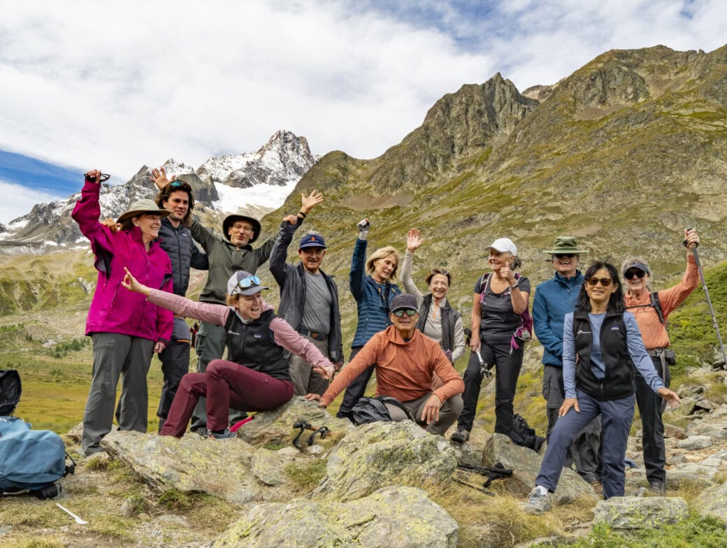 A group of people posing happily on a rocky terrain with mountains in the background, some sporting hiking gear and holding walking sticks, all enjoying the crisp Autumn air as they admire Mont Blanc's majestic beauty.