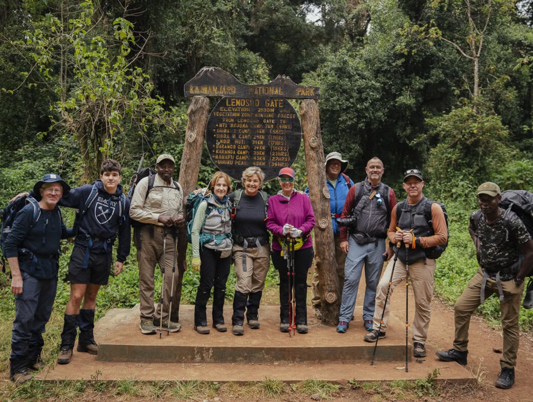 A group of hikers, ready to embark on their adventure, poses before the Kilimanjaro National Park sign at Lemosho Gate, surrounded by dense forest. Perfect for those following a beginner's guide to climbing Mount Kilimanjaro.