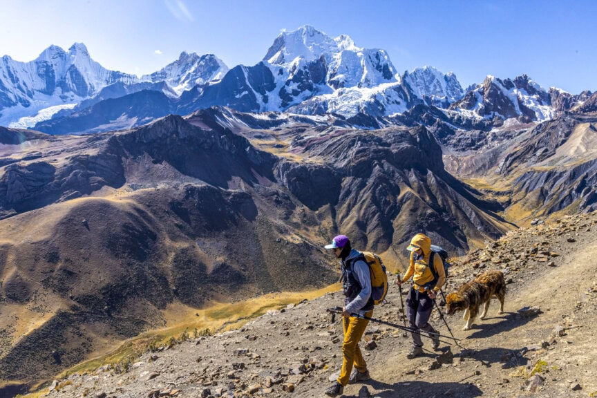 Two hikers with walking sticks and a dog trek a mountain trail in the breathtaking Peruvian Andes, snow-capped peaks soaring in the background under a clear blue sky.