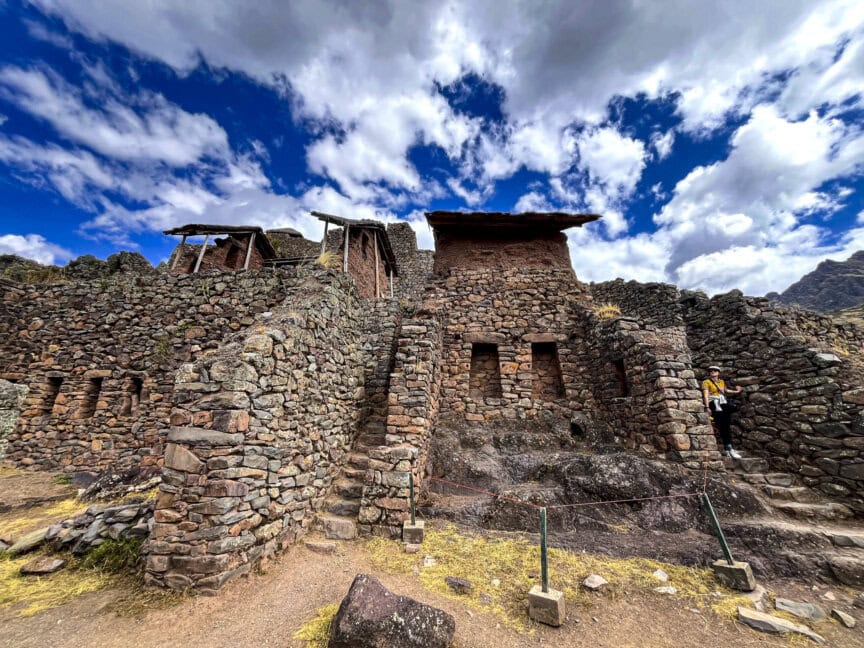 Amid the ancient stone ruins under a partly cloudy sky, a lone hiker stands near the structure on the right, offering a glimpse into the mysteries of the Peruvian Andes.