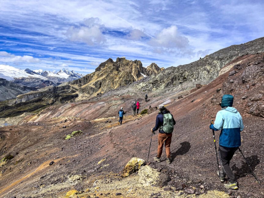 Hikers with trekking poles navigate a rocky mountain path under a partly cloudy sky in the breathtaking Peruvian Andes. Jagged peaks and snow-capped mountains rise majestically in the background, offering an unforgettable hiking experience.