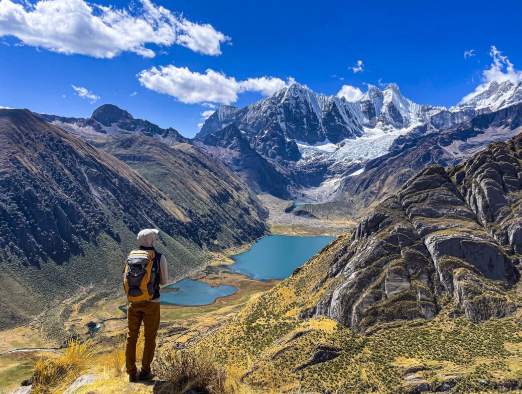 A person with a backpack stands on a grassy hill in the Peruvian Andes, overlooking a valley with two lakes and snow-capped mountains under a clear blue sky.