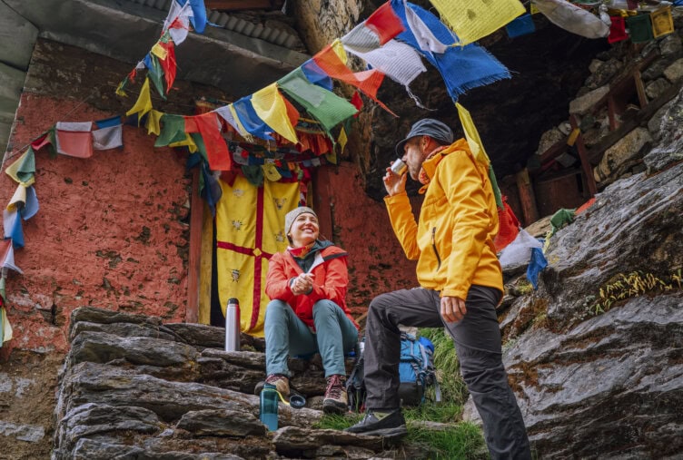 Two people in outdoor gear sit on stone steps near a rustic building adorned with colorful prayer flags, one drinking from a cup, acclimatizing to the serene environment of Nepal, all surrounded by lush greenery.