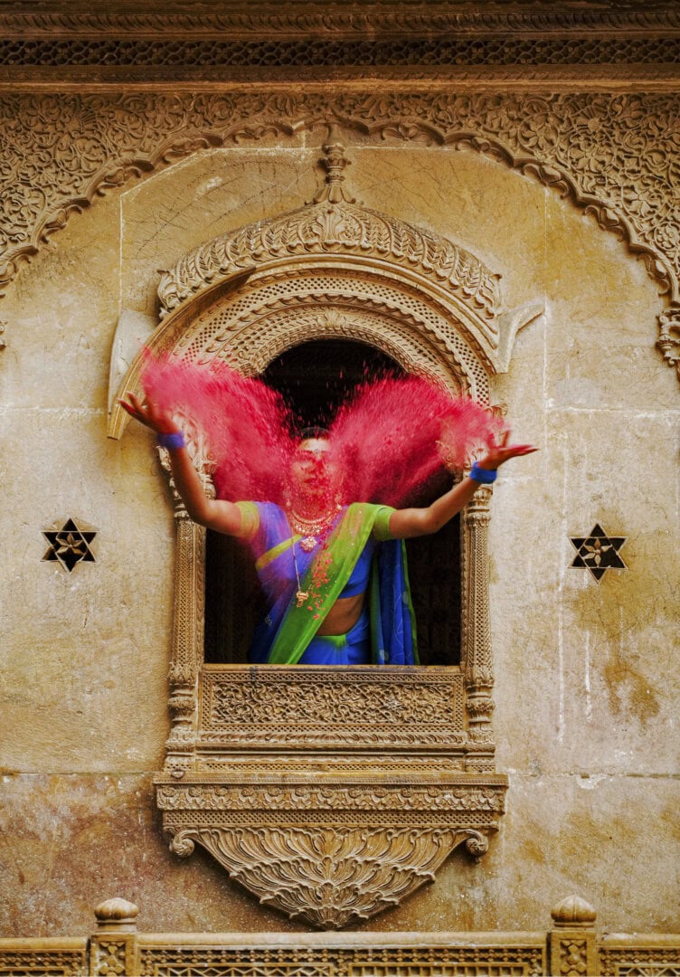 A person in traditional attire throws pink powder from a carved stone balcony, capturing the vibrant spirit of Rajasthan, India.