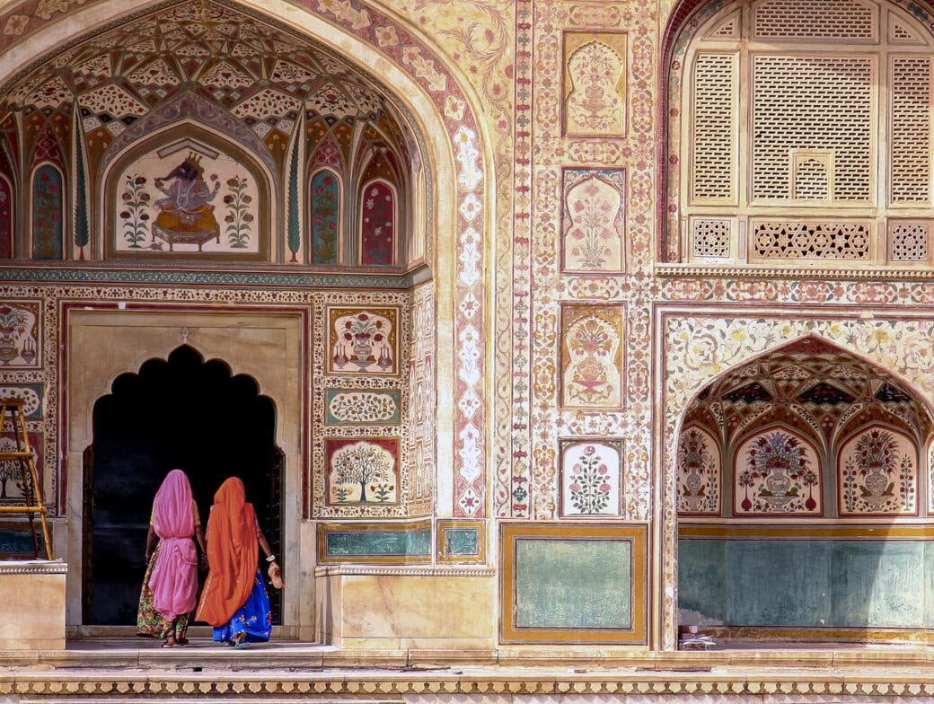 Two people in colorful attire enter an ornately decorated historic building in Rajasthan, graced with detailed floral patterns and arches, offering a glimpse into India's vibrant heritage.