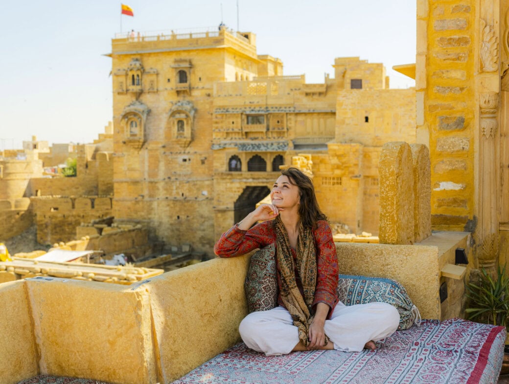 A person sitting cross-legged on a cushioned terrace in Rajasthan, looking up thoughtfully. Historic sandstone buildings rise majestically in the background under a clear sky, making this moment feel like it belongs in a beginner's guide to India.