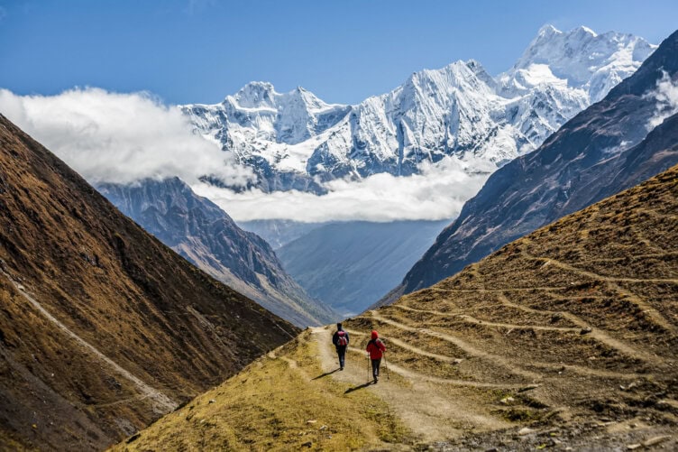 Two people hiking on a dirt trail between steep hills, with snow-covered mountains and a layer of clouds in the background, are training for their upcoming expedition in Nepal.
