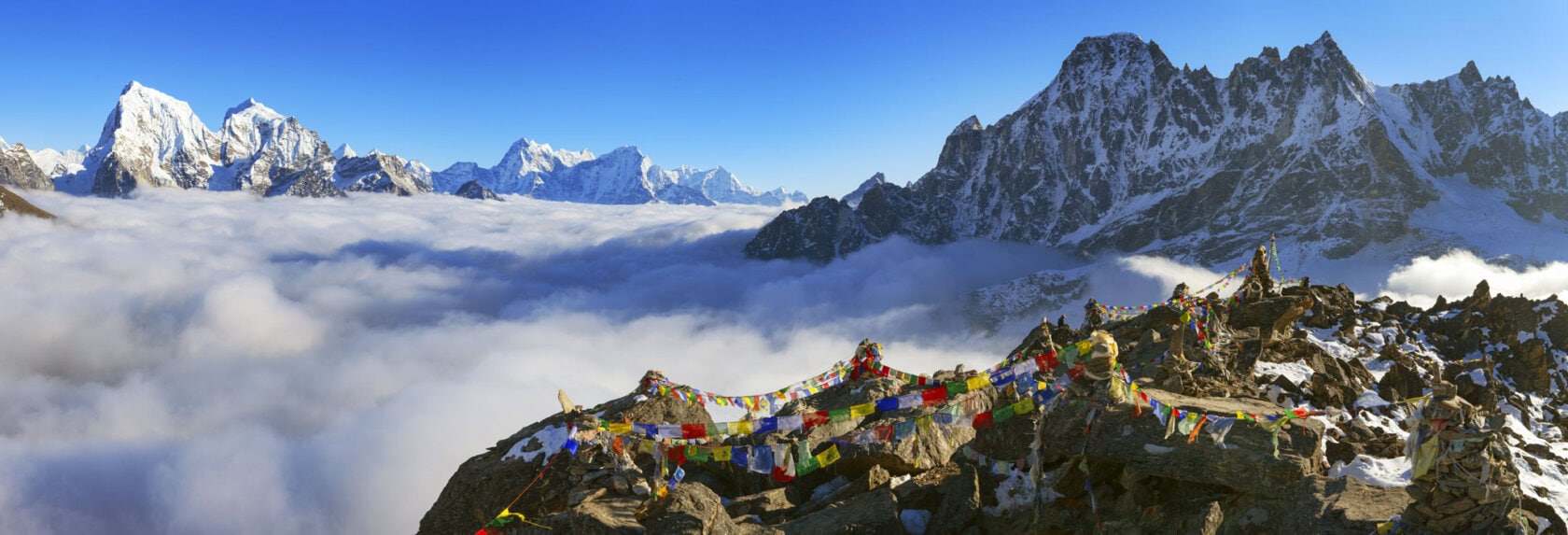 A stunning mountain range with snow-covered peaks rises above the clouds, while vibrant prayer flags flutter in the rocky foreground, painting a serene picture of Nepal's majestic landscape.