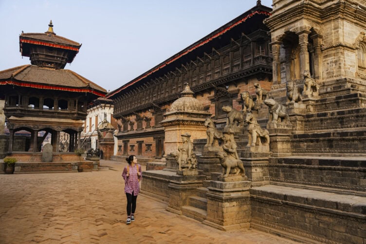 A person walks through a historic plaza in Nepal, lined with stone structures and sculptures, featuring traditional architecture and a reddish-brown brick pathway.