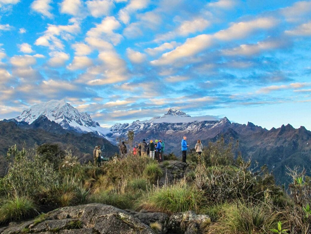 Hikers stand on a grassy hilltop with snow-capped mountains and a partly cloudy sky in the background, reminiscent of the breathtaking views seen when hiking the Inca Trail.