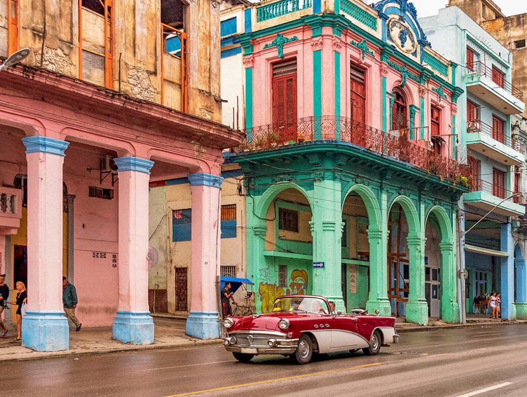 A classic car glides through the vibrant streets of Cuba, past colorful, weathered buildings with arches.