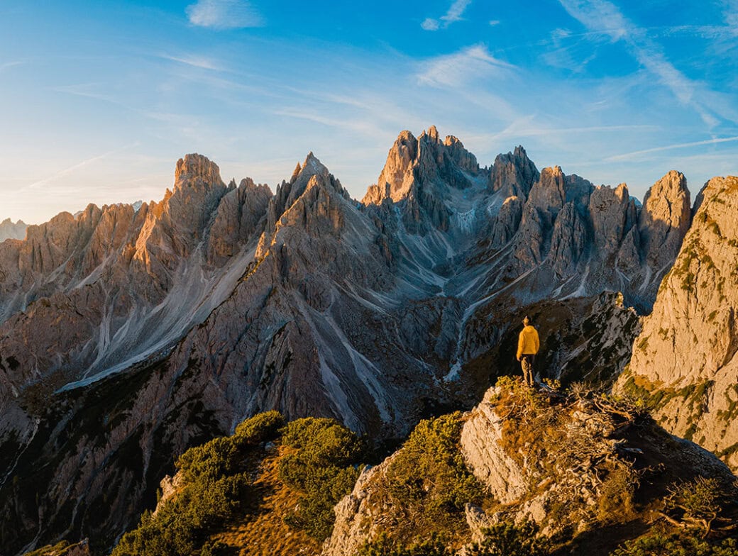 A solo traveler in a yellow jacket stands on a rocky hill, overlooking a rugged mountain range under the clear blue sky—an inspiring scene for those who seek unforgettable travel experiences.