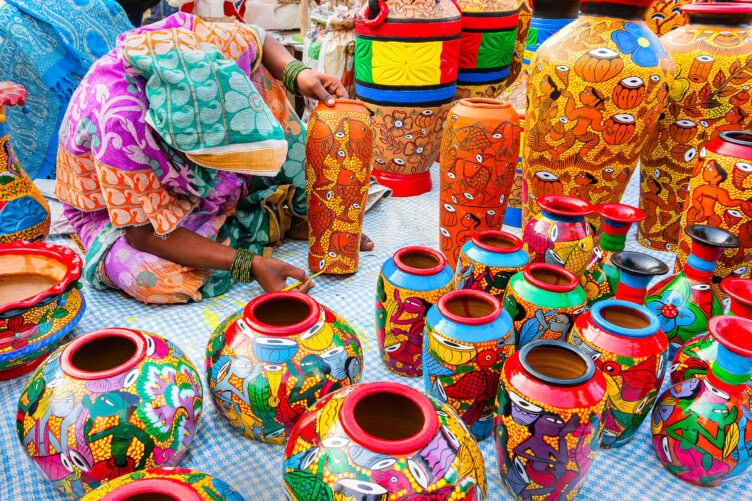 Person in colorful clothing painting vibrant, decorative pottery on a checkered cloth. Various painted vases and pots in diverse patterns and colors are displayed around them—a perfect glimpse into things to do while traveling in India.
