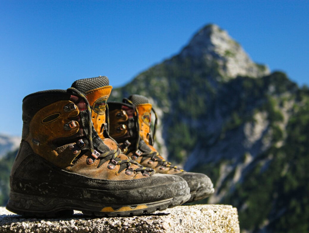 A pair of well-chosen hiking boots rests on a stone ledge, their wear telling tales of adventure. In the background, a mountain peak rises majestically against the clear blue sky, reminding every hiker of nature's call and the expert tips needed for each journey.