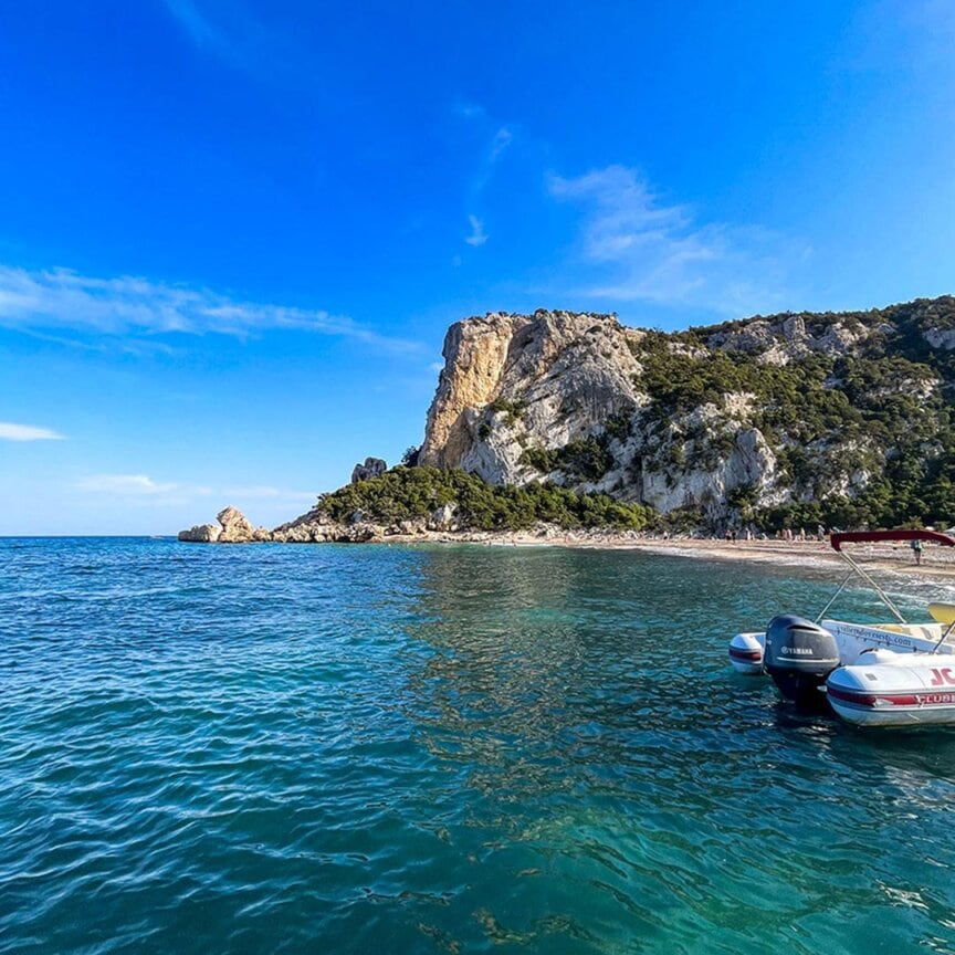 A boat gently docked in the clear turquoise waters of Sardinia, near a rocky shoreline adorned with lush green vegetation under a vibrant blue sky—perfect for adventurers drawn to hiking trails that weave through this Mediterranean paradise.