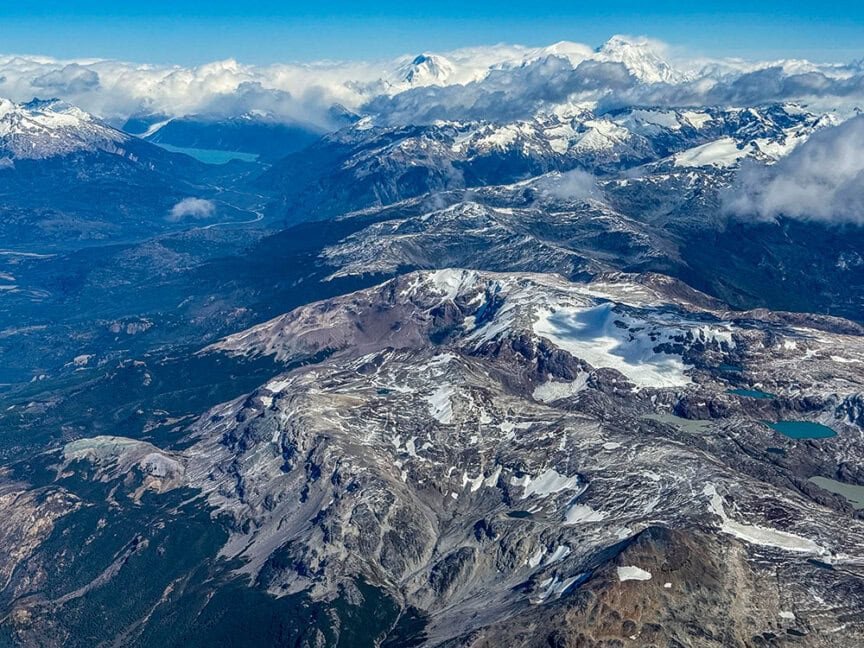 Aerial view of rugged, snow-capped mountains with scattered clouds, glaciers, and alpine lakes under a blue sky—the perfect setting for an unforgettable hiking tour.