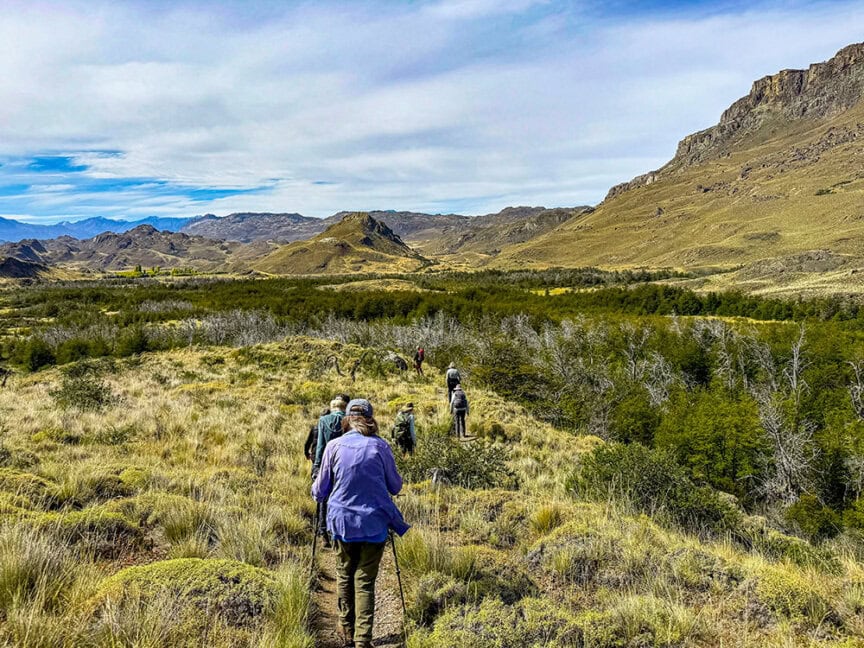 A group of travelers enjoys a hike along a trail through grassy terrain, with mountains and scattered trees in the background under a partly cloudy sky.
