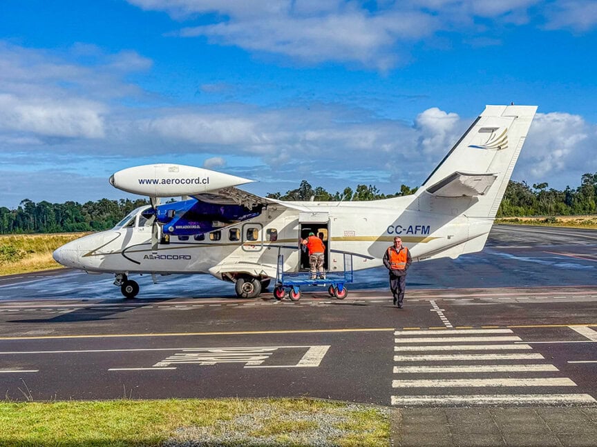 A small white twin-engine airplane with cargo bay open is parked on a runway, as two workers unload gear—perhaps for a traveler's trip—with one standing nearby.