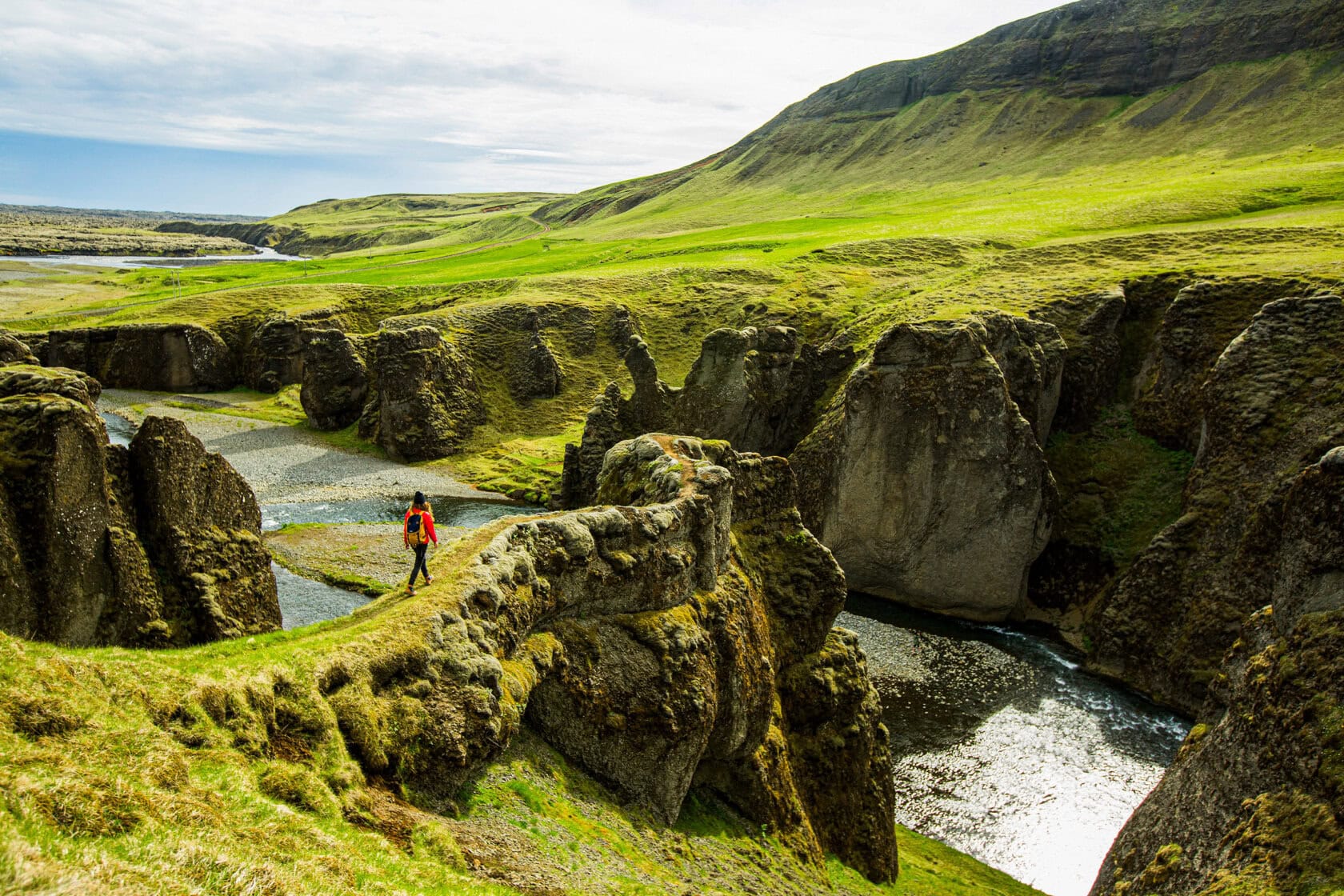 A traveler in an orange jacket hikes along a rocky, grassy path overlooking a river and cliffs in a broad, green valley.