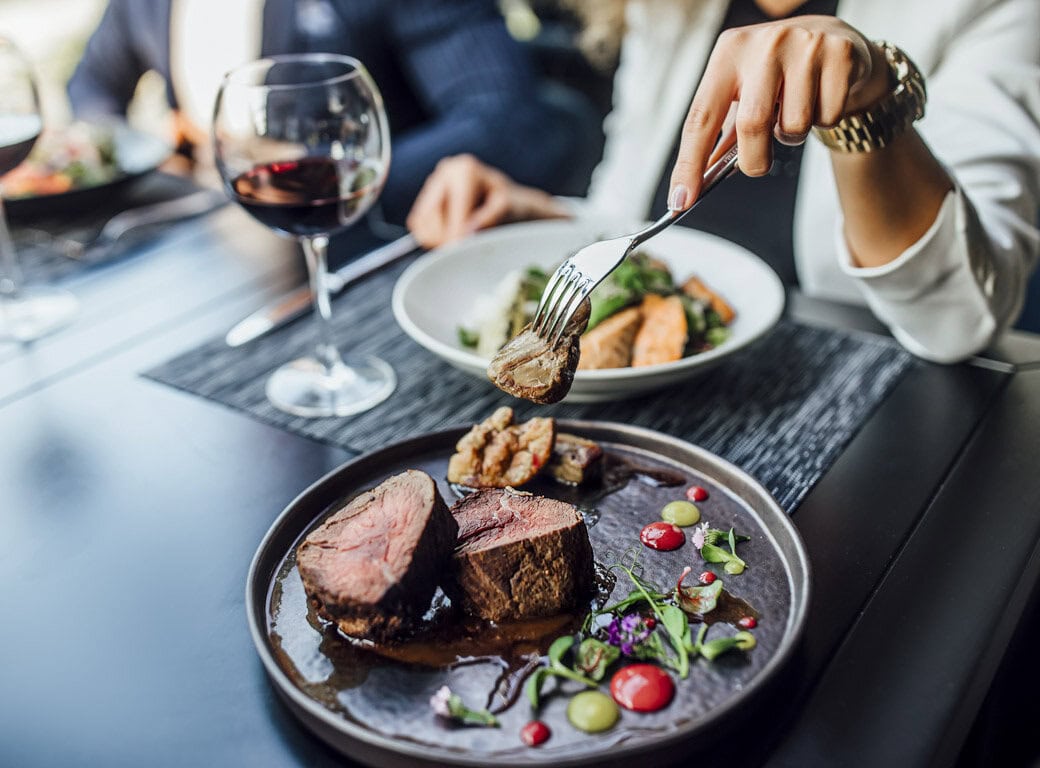 A person holds a fork with a bite of steak over a plate with sliced steak, garnish, and sauce—perfect for savoring local flavors in Juneau. Wine and another dish are visible, offering delicious things to do while you travel.