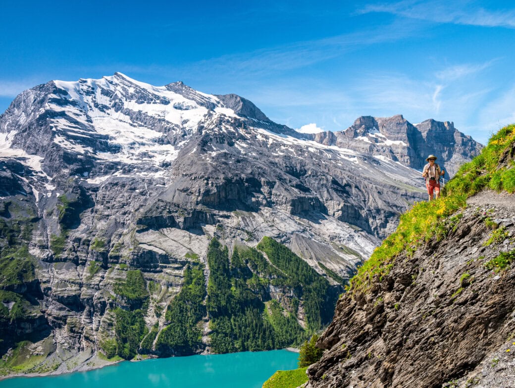A person enjoys hiking alps on a mountain trail overlooking turquoise water, with snow-capped peaks and rocky cliffs in the background under a clear blue sky.