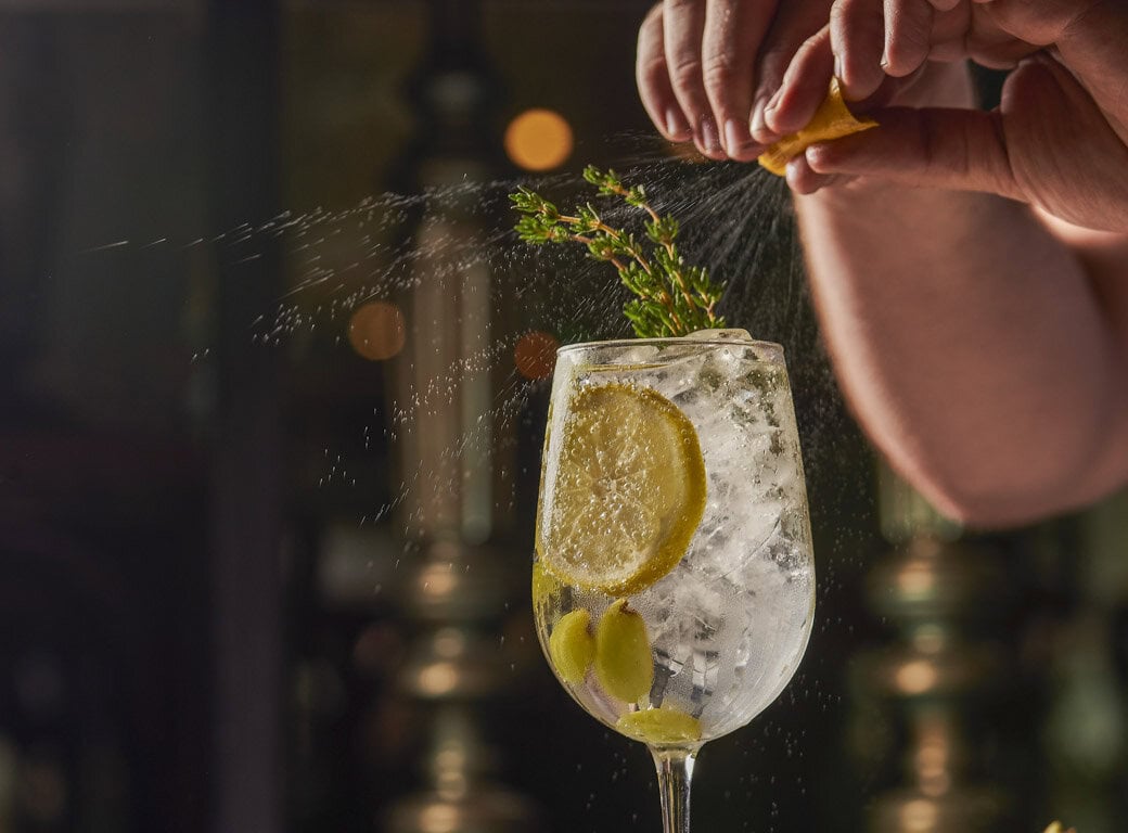 A hand squeezes a lemon peel over a cocktail glass filled with ice, lemon slices, olives, and herbs—a refreshing sip inspired by travel adventures in Juneau and the perfect addition to your list of things to do.