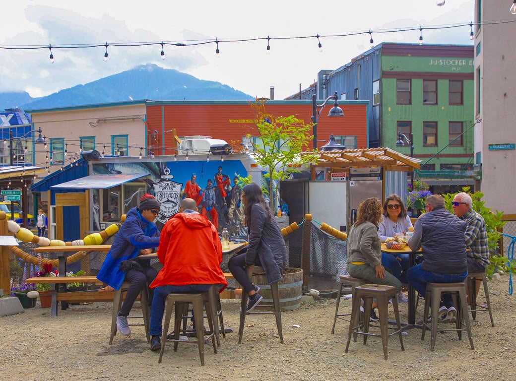 People sit at outdoor tables eating and talking in a casual setting surrounded by colorful buildings, string lights, and mountains—a perfect Juneau spot for relaxing and discovering new things to do during your travel adventure.