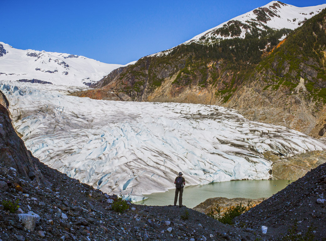 A person stands on rocky terrain overlooking a glacier and a small lake, surrounded by snow-capped mountains under a clear blue sky—one of the top attractions and must-see things to do in Juneau.