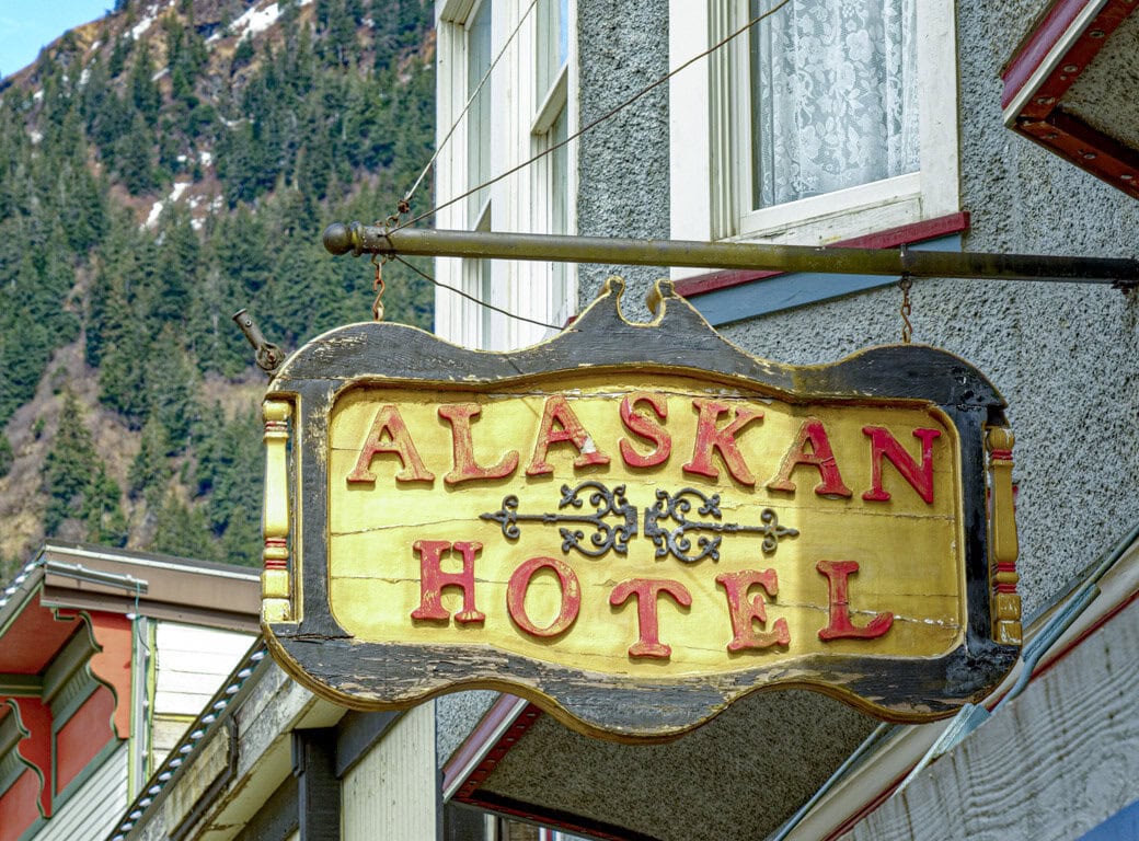 A wooden sign reading "Alaskan Hotel" hangs outside a building in Juneau, with a forested mountain in the background—perfect for travelers exploring things to do in this scenic Alaskan city.