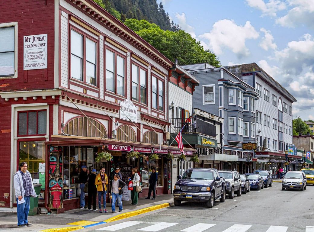 A street scene in Juneau with historic wooden storefronts, parked cars, pedestrians, and shops—including Mt Juneau Trading Post—offers a glimpse of small-town charm and highlights unique things to do for travel enthusiasts on a cloudy day.