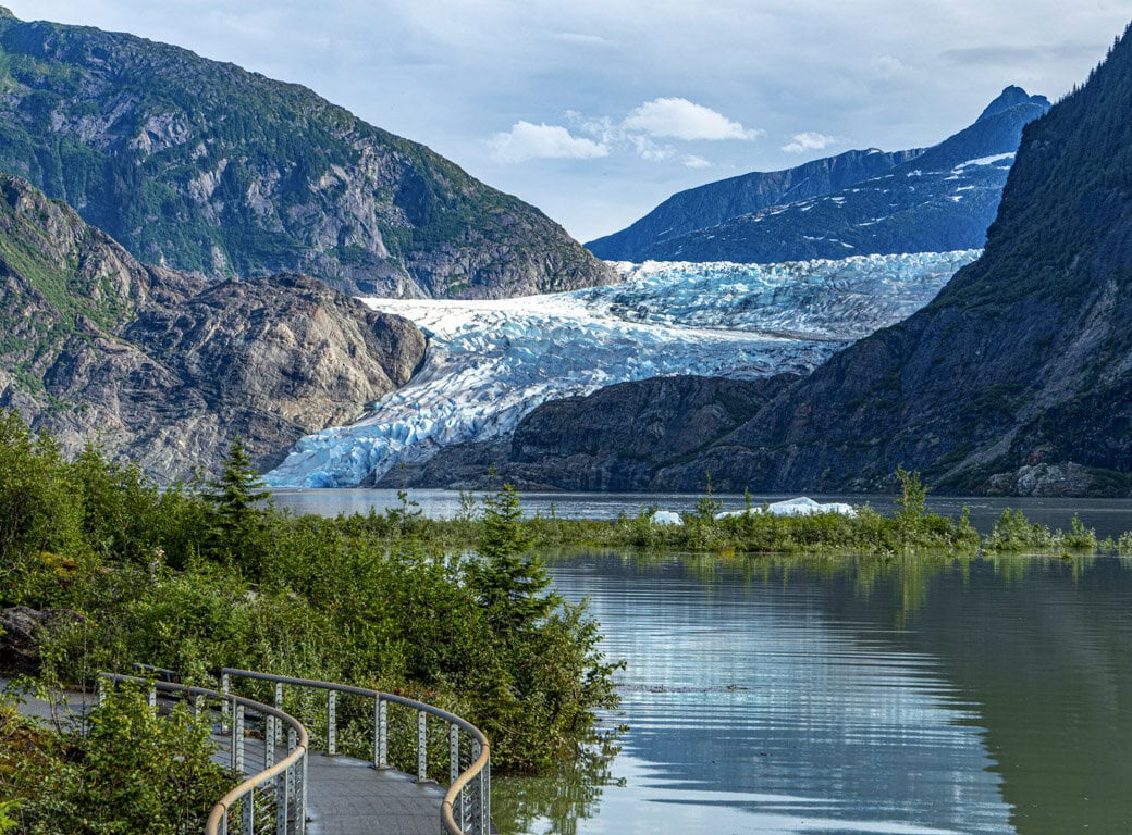 A glacier stretches between mountains above a lake, with a wooden walkway winding through green vegetation in the foreground—one of the unforgettable things to do in Juneau.
