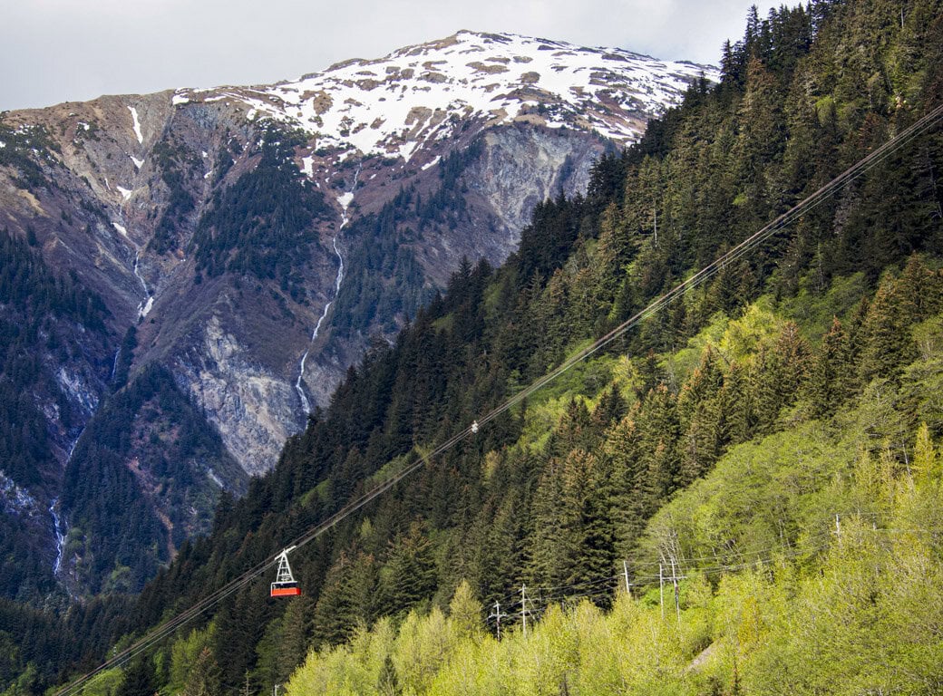 A red cable car travels up a forested mountainside in Juneau, with a snow-capped peak visible in the background—one of the top attractions and unforgettable things to do in the area.